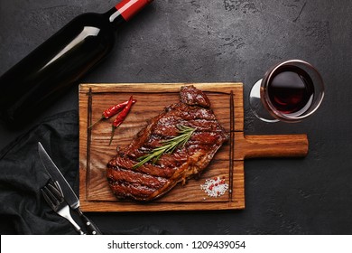 Striploin Grilled Beef Steak Served With Rosemary, Salt And Peppercorns On Wooden Board With Glass Of Wine, Bottle Of Wine, Fork And Knife. Overhead View