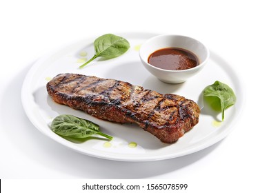 Striploin Steak With Sauce Close Up. Grilled Beef With Basil Leaves Isolated On White Background. Barbeque Meat, Roasted Pork Served On Plate Composition. Traditional New York Strip Steak