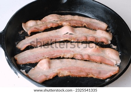 Strips of uncooked bacon sizzling in cast iron frying pan