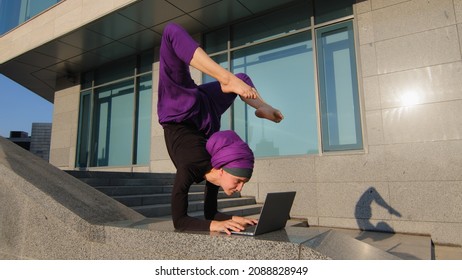 Strong acrobat business woman funny muslim girl in hijab stands on hands in acrobatic yoga pose asana balance handstand in city outdoors typing laptop working remotely online keep legs in air position