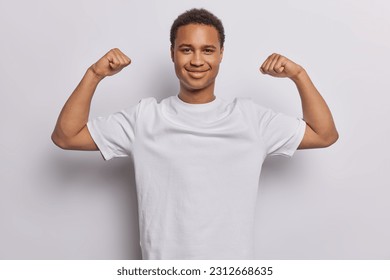 Un hombre fuerte de África levanta los brazos y muestra a los músculos de la bicicleta posando con una camiseta casual sobre fondo blanco. Un tipo sonriente que está en gran condición física haciendo ejercicio en el gimnasio regañando los logros