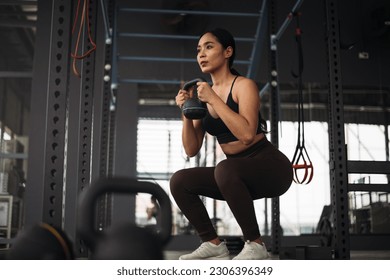 Strong Asian woman doing exercise with kettlebell at cross fit gym. Athlete female wearing sportswear doing squat workout on grey gym background with weight and dumbbell equipment. Healthy lifestyle. - Powered by Shutterstock