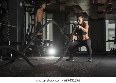 Strong athlete woman wearing a black sports bra and long tights in a dark gym with a muscular male in the background  using  battle ropes for exercise