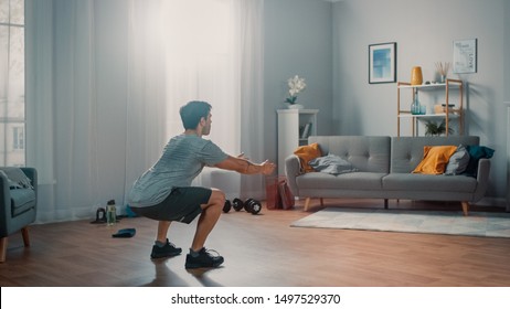 Strong Athletic Fit Man in T-shirt and Shorts is Doing Squat Exercises at Home in His Spacious and Bright Apartment with Minimalistic Interior.