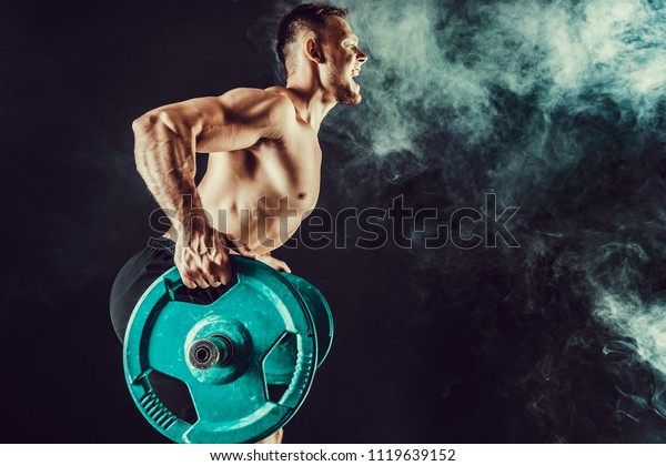 強い運動選手が筋肉を増やしてボディービルをする 暗い背景に煙のあるバーベルを持つ背中の筋肉の運動をする筋肉質のボディビルダーのハンサムな男性 強さと動機 の写真素材 今すぐ編集