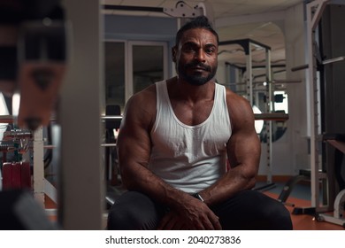 A Strong Athletic Man In A White T-shirt And Black Sweatpants Sits In The Gym On The Simulator. High Quality Photo