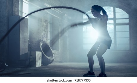 Strong Athletic Woman Exercises with Battle Ropes as Part of Her Fitness Gym Workout Routine. She's Covered in Sweat and Training Takes Place in a Abandoned Factory Remodeled into Gym.