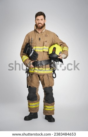 Strong, bearded fireman posing to camera with yellow protective helmet. Front view of happy male firefighter keeping hand on leather belt, while looking at camera, on gray background. Concept of work.