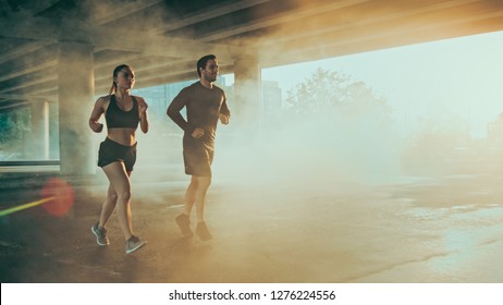 Strong and Beautiful Athletic Fitness Couple in Workout Clothes Jogging in a Street Filled With Steam on a Sunny Day. They are Running in an Urban Environment Under a bridge.