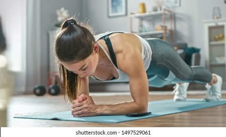 Strong Beautiful Fitness Girl in Athletic Workout Clothes is Doing a Plank Exercise While Using a Stopwatch on Her Phone. She is Training at Home in Her Living Room with Cozy Interior.