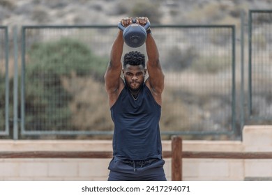 Strong black sportsman in activewear with Afro hair performing kettlebell swing exercise during intense fitness training on sports ground against net fence - Powered by Shutterstock