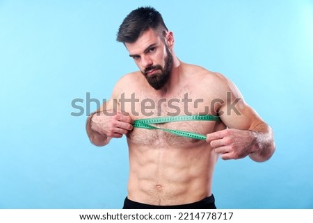 Strong bodybuilder measuring the width of his chest, putting a measuring tape over his abdomen, blue studio background