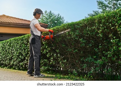 Strong caucasian man in uniform, safety gloves and mask pruning hedge with petrol trimmer. Male gardener shaping green overgrown bushes outdoors.
