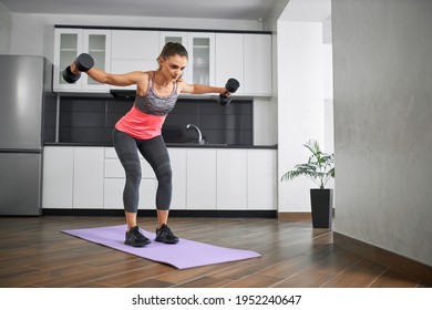 Strong Caucasian Young Woman Raising Dumbbells On Mat In Kitchen. Fit Girl Wearing Sportswear Training Shoulders At Home In Morning. Concept Of Home Workout, Weight Loss.