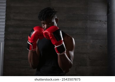 strong confident african american boxer in red gloves stands in a defensive stance near a punching bag, athlete in red gloves looks at the camera on a black background - Powered by Shutterstock