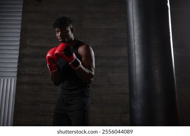 strong confident african american boxer in red gloves stands in a defensive stance near a punching bag, athlete in red gloves looks at the camera on a black background - Powered by Shutterstock