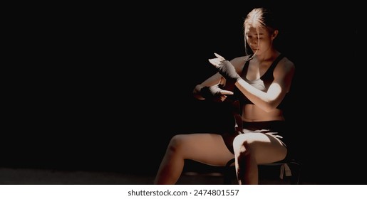 Strong and confident young Asian Muay Thai boxer woman sitting and wrapping hands with sport bandage on black background, copy space banner size - Powered by Shutterstock