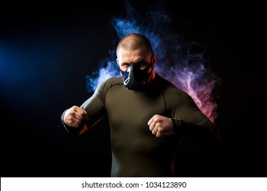 A strong dark-haired sportman wrestlers in a green sports shirt and training mask  posing against a blue and red vape smoke background on a black isolated  - Powered by Shutterstock