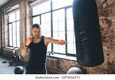 Strong determined male boxer with hands wrapped in tape for protection and looking away while preparing to attack punching bag in foreground in gym with equipment - Powered by Shutterstock