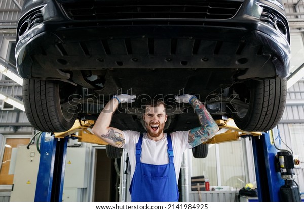 Strong excited young male professional\
technician mechanic man wears denim blue overalls white t-shirt\
stand near car lift posing like carry automobile work in vehicle\
repair shop workshop\
indoors.