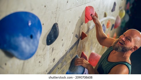 Strong Experienced Rock Climber Practicing Solo Climbing On Bouldering Wall In Gym. Man Exercising At Indoor Fitness Facility, Doing Extreme Sport For His Healthy Lifestyle Training. Close Up Portrait