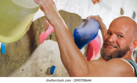 Strong Experienced Rock Climber Practicing Solo Climbing On Bouldering Wall In Gym. Man Exercising At Indoor Fitness Facility, Doing Extreme Sport For His Healthy Lifestyle Training. Close Up Portrait