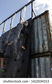 Strong And Fast Girl Climbing To The Top Of The Metal Wall Through The Waterfall. Sun Shining, Blue Sky, Summer War Day. Man And Woman Compete In A Race With Barriers, Interesting Activities.