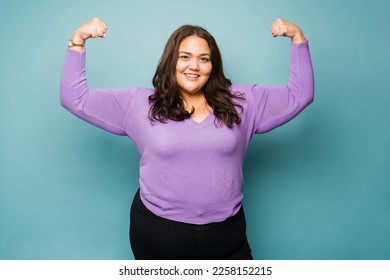 Strong fat hispanic woman feeling powerful making bicep curl looking happy smiling against a studio background - Shutterstock ID 2258152215