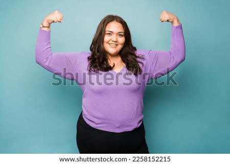 Strong fat hispanic woman feeling powerful making bicep curl looking happy smiling against a studio background