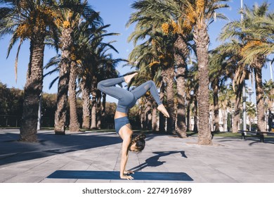 Strong female doing handstand while practice body balance at city streets with tropical palm trees, Asian woman in tracksuit training and recreating during hatha workout reaching healthy lifestyle