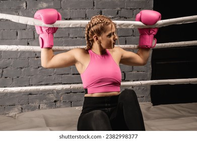 Strong female fighter in pink boxing gloves is tired and leaning on the ropes in the ring and resting in the gym - Powered by Shutterstock