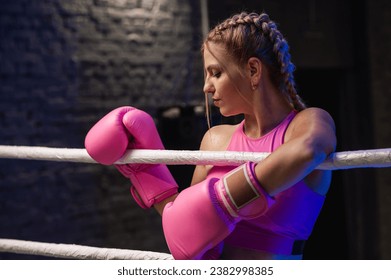 Strong female fighter in pink boxing gloves is tired and leaning on the ropes in the ring and resting in the gym - Powered by Shutterstock