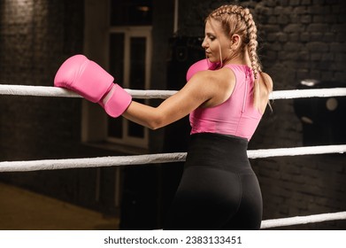 Strong female fighter in pink boxing gloves is tired and leaning on the ropes in the ring and resting in the gym - Powered by Shutterstock