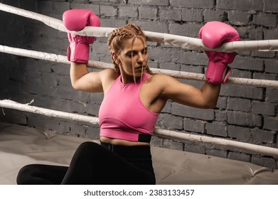 Strong female fighter in pink boxing gloves is tired and leaning on the ropes in the ring and resting in the gym - Powered by Shutterstock