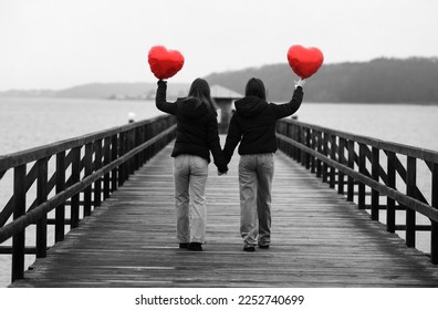                      Strong female friendship. Back view of two teenage girls best friends holding hands with heart shaped balloons in hands while walking on a bridge outdoors. black and white photo.  - Shutterstock ID 2252740699