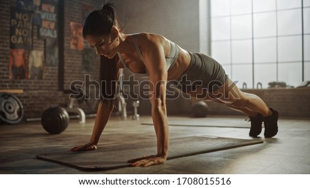 Strong and Fit Athletic Woman in Sport Top and Shorts is Doing Push Up Exercises in a Loft Style Industrial Gym with Motivational Posters. It's Part of Her  Fitness Training Workout. Warm Light. Foto stock © 