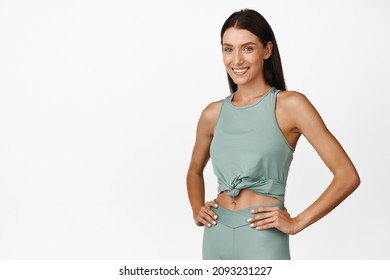 Strong and fit female athlete standing in sprotswear, hold hands on waist and smiling confident, workout in gym, concept of training and sport, white background