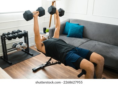 Strong fit man in his 20s with a home gym doing bench press exercises with dumbbell weights - Shutterstock ID 2268627955