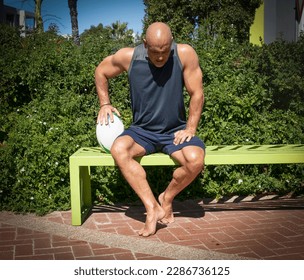 Strong and fit rugby player sitting on a green bench holding a rugby ball and looking down. - Powered by Shutterstock