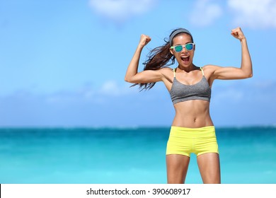 Strong fitness funny woman in neon blue wayfarer sunglasses on beach showing off muscular arms flexing biceps for fun. Fit girl in sportswear after running strength training workout winning in power.