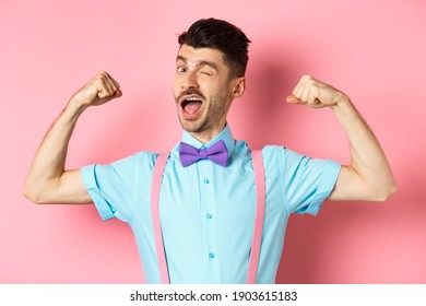 Strong And Funny Guy With French Moustache, Flexing Biceps And Winking At Camera, Show-off His Strengths, Standing Over Pink Background