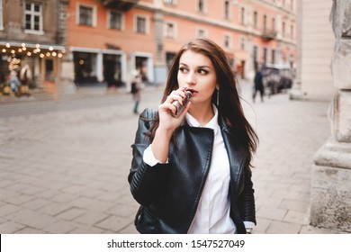 Strong girl in a black leather jacket smokes an electronic cigarette on a city street - Powered by Shutterstock