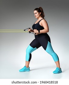 Strong girl working with resistance band. Photo of model with curvy figure in fashionable sportswear on grey background. Sports motivation and healthy lifestyle