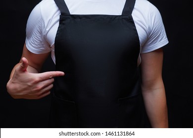 Крепкий туловище парня в черном фартуке, показывающем пальцем к центру фартука. Макет концепции шеф-повара или официант. Темный фон