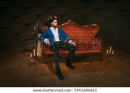 Strong handsome confident man with ram horns on head sitting on red vintage sofa in dark room. Enchanted prince beast monster. Blue jacket kaftan vintage white shirt. Stubborn male king fashion model