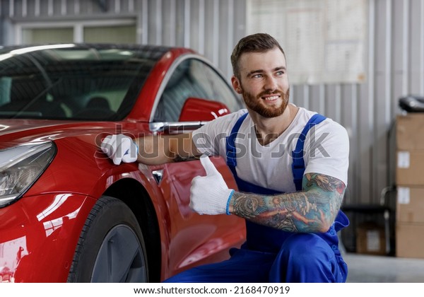 Strong happy minded young professional technician\
car mechanic man in denim blue overalls white t-shirt gloves check\
wheel show thumb up look aside work in modern vehicle repair shop\
workshop indoor