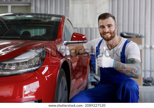 Strong happy satisfied young professional\
technician car mechanic man 20s in denim blue overalls white\
t-shirt gloves check wheel show thumb up work in light modern\
vehicle repair shop workshop\
indoor