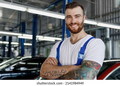 Strong happy young technician car mechanic man in denim blue overalls white t-shirt look camera hold hands crossed folded work in light modern vehicle repair shop workshop indoors Tattoo translate fun
