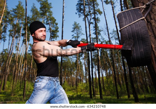 Strong healthy adult ripped\
man with big muscles hitting car tyre with big hammer. Outdoors\
workout, sports, power fitness, willpower, endurance exercises\
concept