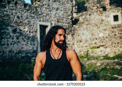 Strong Healthy Muscular Man With Long Wavy Hair And Beard At Ancient Wall Background. Mens Style And Wellness Concept. Strong Muscular Handsome Man Walking Outside